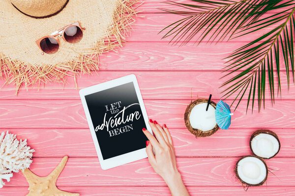 cropped shot of woman using tablet with "let the adventure begin" lettering on pink wooden surface with coconut cocktail and straw hat 
