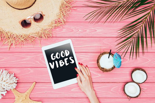 cropped shot of woman using tablet with "good vibes" inspection on pink wooden surface with coconut cocktail and straw hat