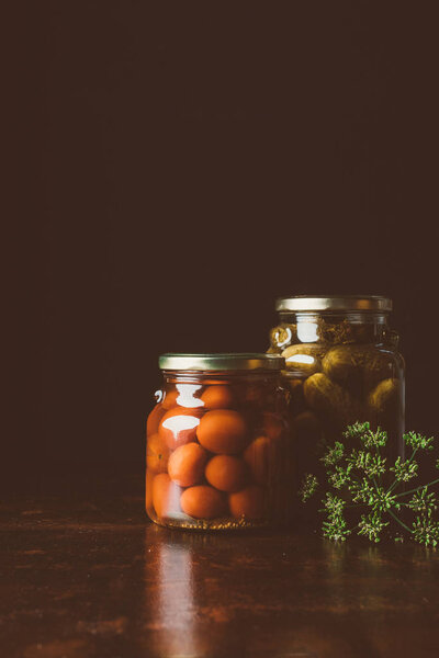glass jars with preserved tomatoes and cucumbers on wooden table in dark kitchen 