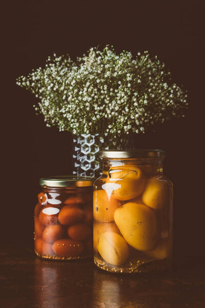 glass jars with preserved tomatoes and flowers on wooden table in dark kitchen 