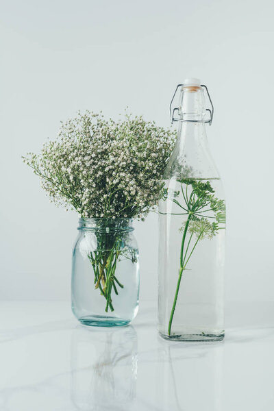 glass bottle with water and dill and glass jar with flowers on white table