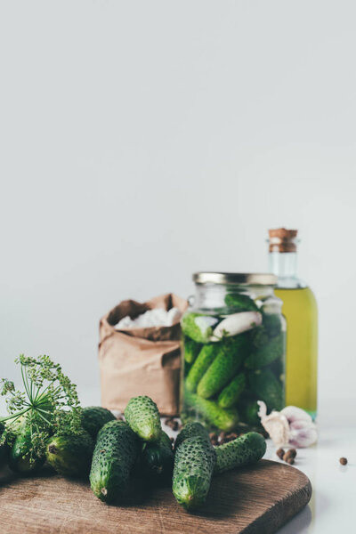 fresh cucumbers on cutting board on table with preserved cucumbers in glass jar on background