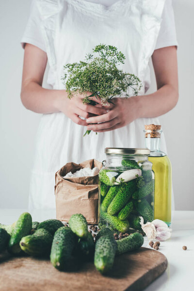cropped image of woman preparing preserved cucumbers and holding dill at kitchen