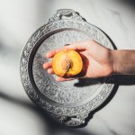 Tiro cortado de mulher segurando peça de pêssego na bandeja de metal na superfície de mármore leve