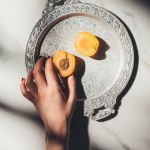 Abgeschnittene Aufnahme einer Frau mit Aprikosenstück auf Metalltablett auf heller Marmoroberfläche