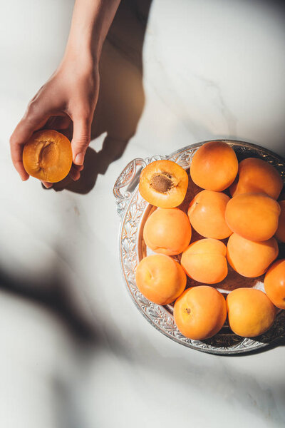 partial view of woman holding piece of apricot with metal tray full of ripe apricots near by on light marble surface