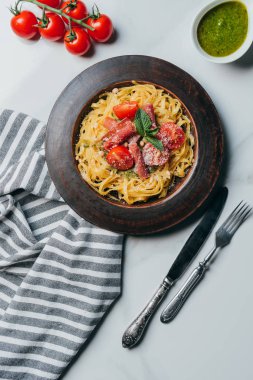Yukarıdan görünüm makarna ile nane yaprakları, jamon ve kiraz domates plaka mermer masa mutfak havlusu, bıçak, çatal ve pesto kase ile parmesan ile kaplı 
