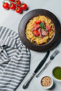 Yukarıdan görünüm makarna ile nane yaprakları, jamon ve kiraz domates kapta çam fıstığı ve pesto sos plaka mermer masa mutfak havlusu, bıçak, çatal, parmesan ile kaplı 