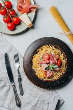 Mutfak havlu, çatal, bıçak ve pişmemiş spagetti ile mermer tabloda makarna, parmesan ve jamon jamon plakaların üstten görünüm 
