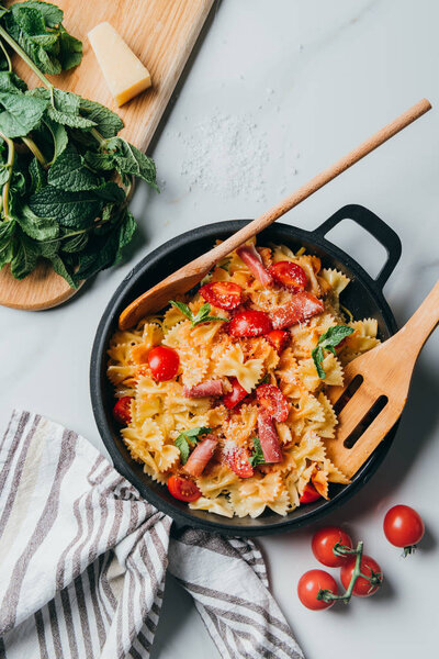 elevated view of pasta with jamon, cherry tomatoes, mint leaves covered by grated parmesan in pan with wooden spatula and spoon on marble table