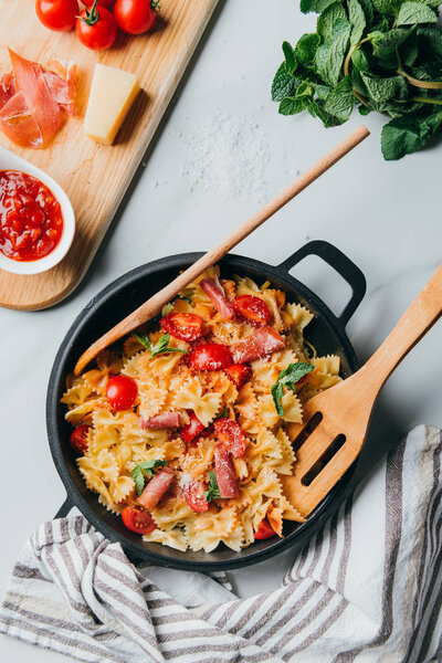 view from above of pasta with jamon, cherry tomatoes, mint leaves covered by grated parmesan in pan with wooden spatula and spoon on marble table