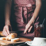 Abgeschnittenes Bild einer Frau in Schürze, die Baguette an Bord schneidet, in der Nähe von Frischkäse und Marmeladenglas am Tisch