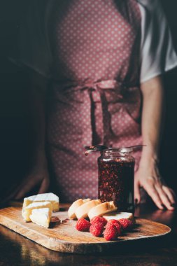 cropped image of woman in apron standing near table with brie, baguette slices, raspberries and jar of jam on cutting board  clipart