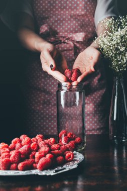 cropped image of woman in apron putting raspberries in jar for making jam  clipart