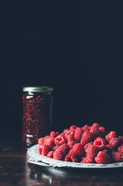 close up view of pile of raspberries in silver tray and jam in jar on black  clipart