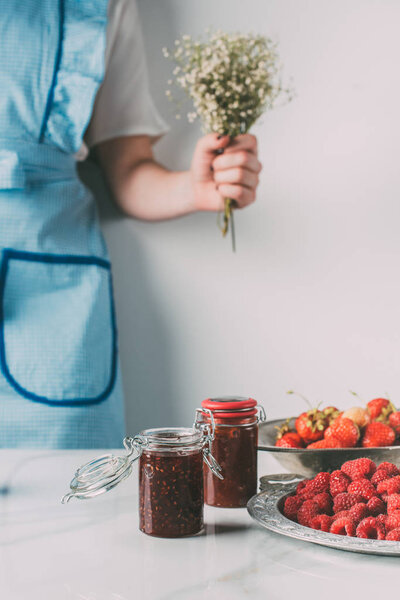 cropped image of woman in apron holding flowers near table with raspberries, strawberries and jars of jam 