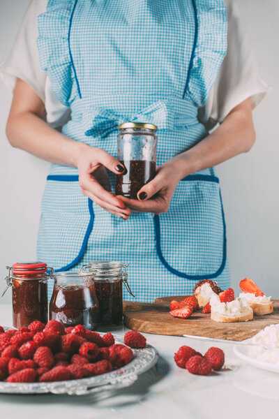 cropped image of woman in apron holding jar with fruit jam at kitchen 