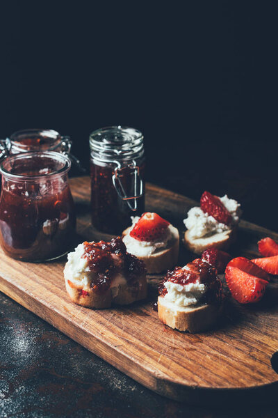 крупным планом банки с вареньем, бутерброды со сливочным сыром, клубничные ломтики и фруктовое варенье на разделочной доске на черном
 