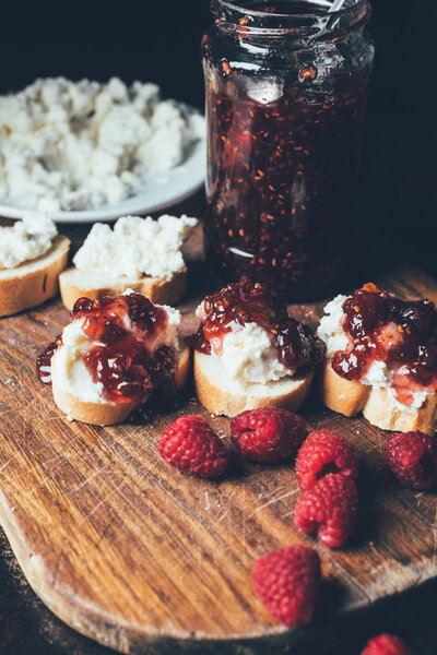 закрытый вид на малину, бутерброды с фруктовым вареньем и сливочным сыром на разделочной доске на черном
 