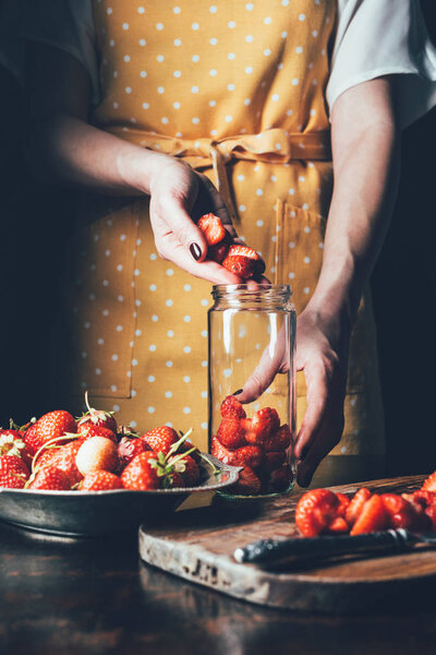 partial view of woman in apron standing at table and putting strawberries in jar 