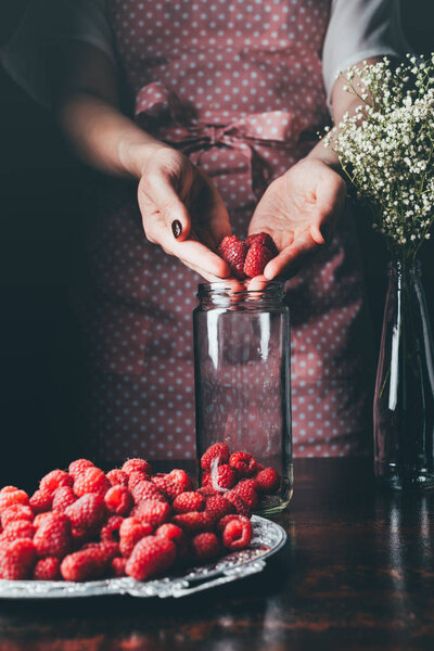 cropped image of woman in apron putting raspberries in jar for making jam 