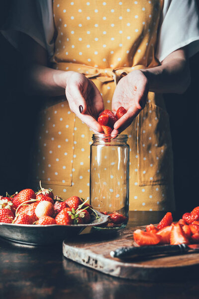 partial view of woman in apron putting strawberries in jar for cooking jam 