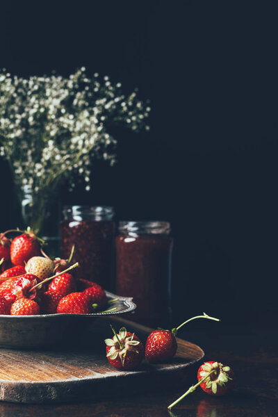 selective focus of strawberries in silver tray, flowers and jam in jars on black 