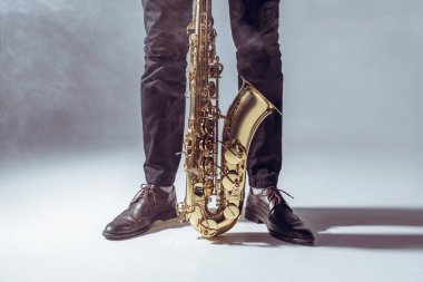 partial view of professional musician standing with saxophone in smoke on grey     clipart