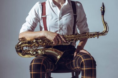 cropped shot of young musician sitting on chair and holding saxophone isolated on grey clipart