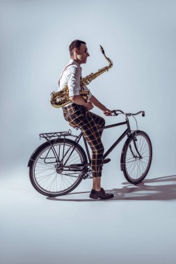 young stylish musician in sunglasses holding saxophone and riding bicycle on grey