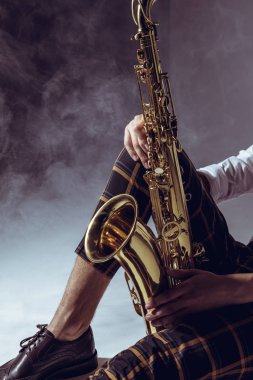 cropped shot of stylish musician holding saxophone in smoke on grey clipart