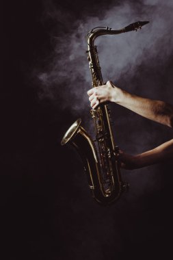 cropped shot of musician holding saxophone in smoke on black clipart