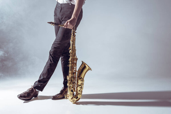 cropped shot of professional musician standing with saxophone in smoke on grey   