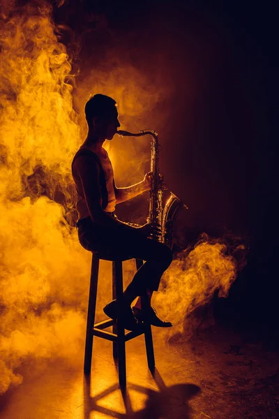 年轻音乐家坐在凳子上玩萨克斯烟雾的剪影 — 图库照片