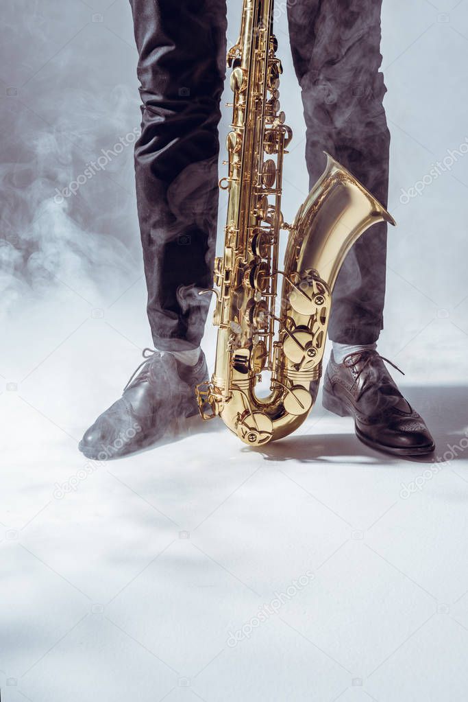 partial view of young musician standing with saxophone in smoke on grey    