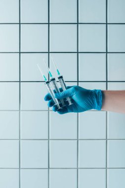 cropped shot of doctor in glove holding syringes in front of tiled white wall clipart