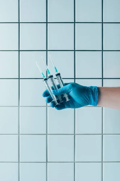 cropped shot of doctor in glove holding syringes in front of tiled white wall
