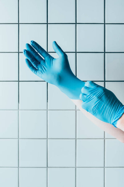 cropped shot of doctor putting on blue rubber gloves in front of tiled white wall