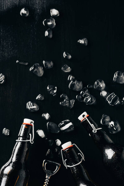 flat lay with bottle opener, glass bottles of beer and ice cubes arranged on dark tabletop