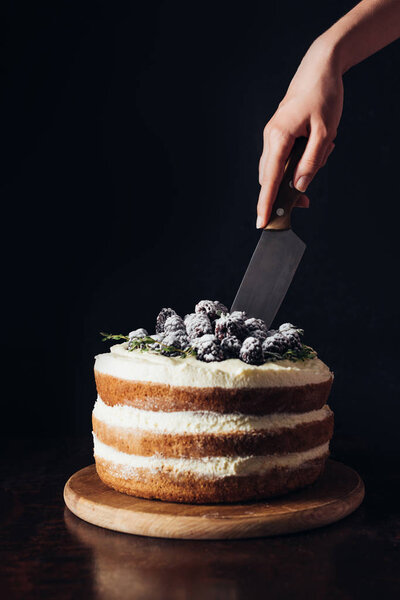 cropped shot of woman cutting delicious blackberry cake on black