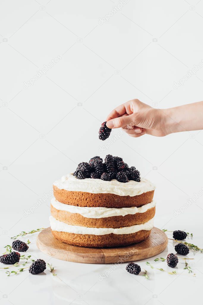 cropped shot of woman decorating tasty blackberry cake on glass stand on white