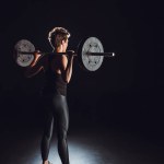 Rear view of young female athlete working out with barbell, black background