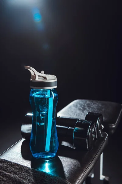 selective focus of sport bottle and dumbbells at gym, black background