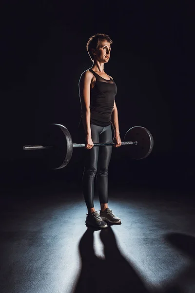 serious sportswoman exercising with barbell at gym, black background