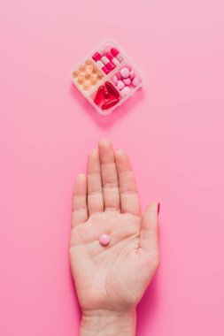 cropped shot of woman holding pink pill on pink surface with plastic container for medicines