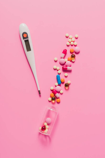 top view of various spilled pills from bottle and electric thermometer on pink