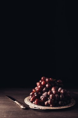 close-up view of fresh ripe red grapes on vintage plate and knife on wooden table on black  clipart