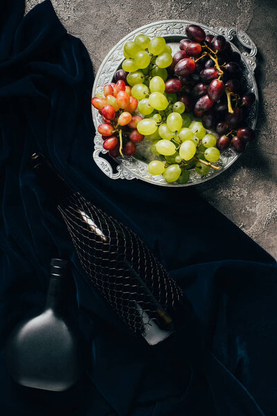 top view of various types of grapes on vintage plate and bottles of wine on dark fabric