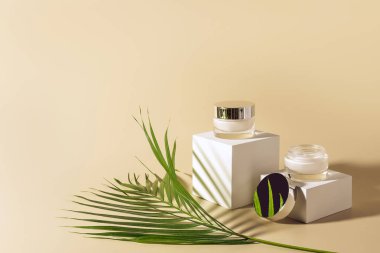 close up view of green palm leaf, facial and body creams in glass jars on white cubes on beige background clipart