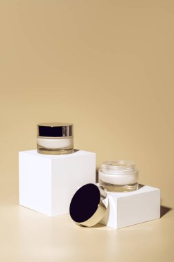 close up view of facial and body creams in glass jars on white cubes on beige background clipart